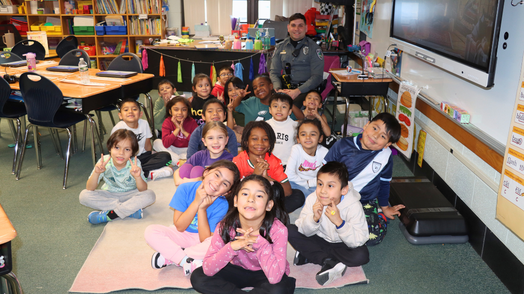 A group of first-grade students sitting on a rug in a classroom with a Officer Dinolfi seated behind them.