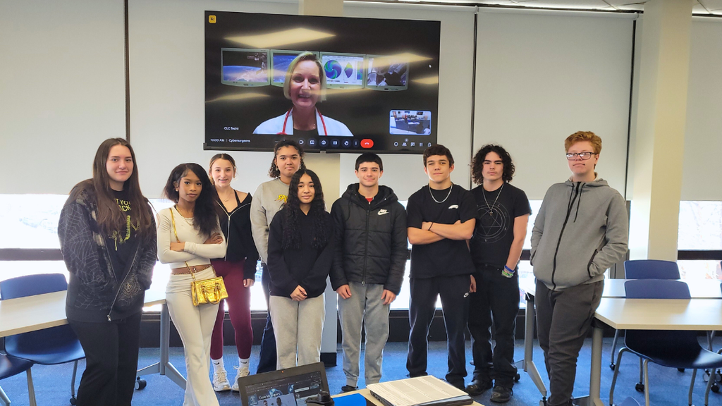 A group of nine students in a classroom, standing in front a screen with a video call, surrounded by tables and chairs.