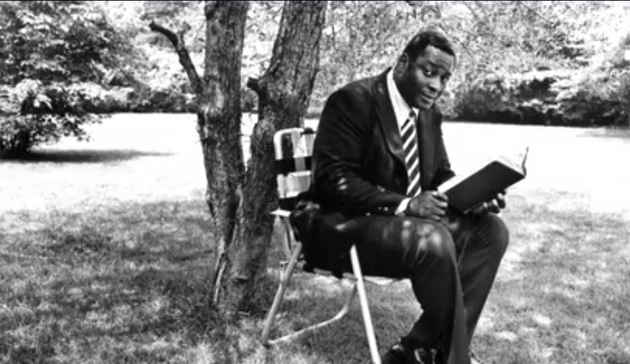 Charles Blockson reads a book in a lawn chair under a tree with shade.