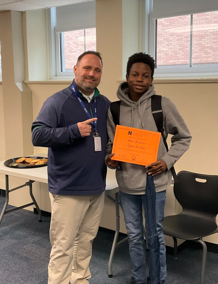 A student is posing with the principal in a classroom after receiving an award. They have the award in their hand.