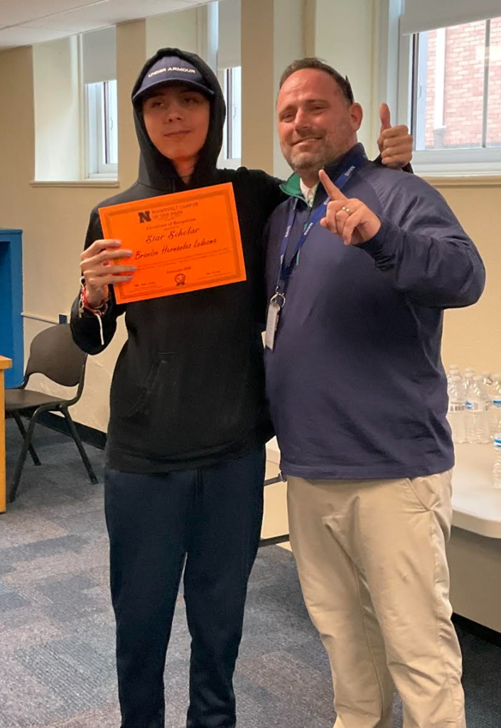A student is posing with the principal in a classroom after receiving an award. They have the award in their hand.
