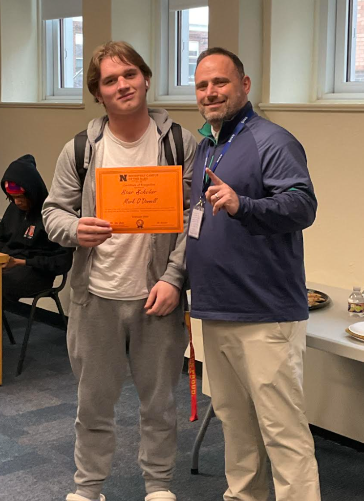 A student is posing with the principal in a classroom after receiving an award. They have the award in their hand.