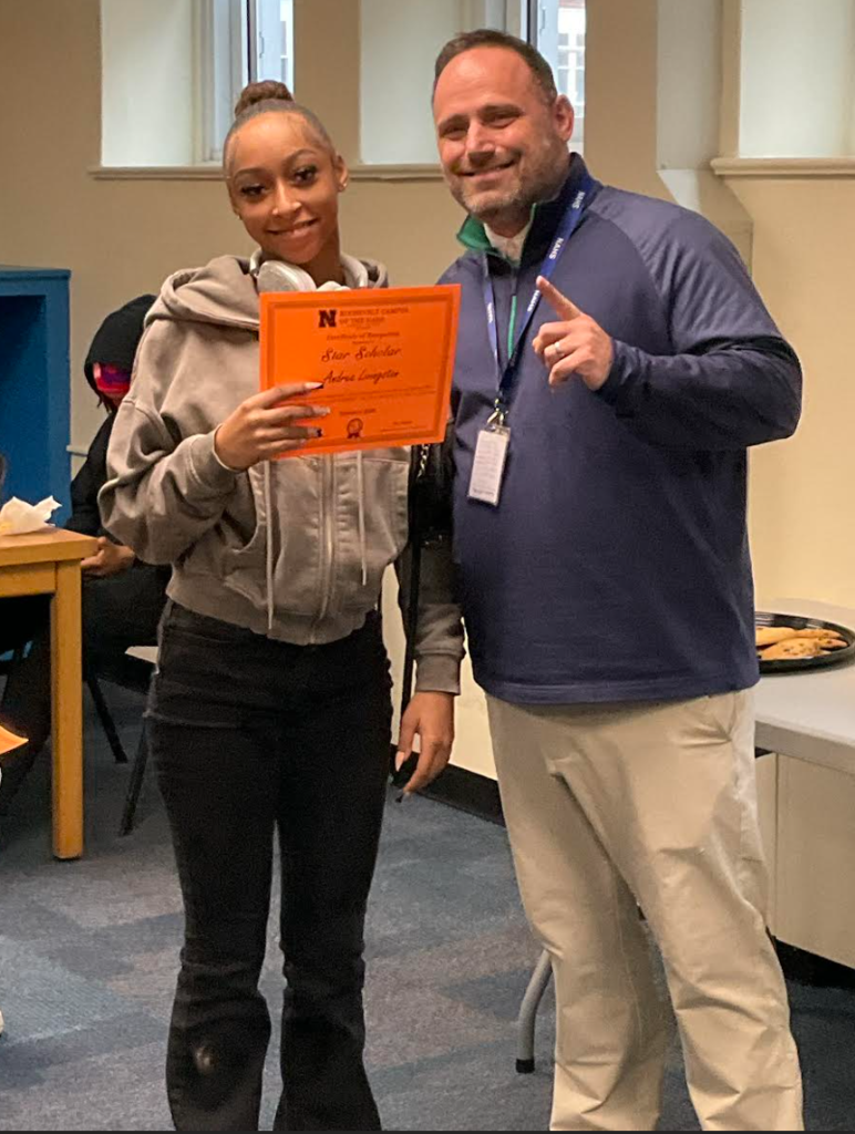 A student is posing with the principal in a classroom after receiving an award. They have the award in their hand.