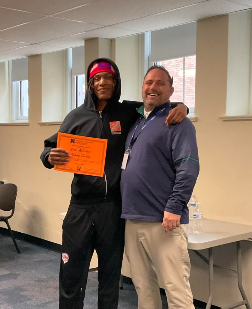 A student is posing with the principal in a classroom after receiving an award. They have the award in their hand.