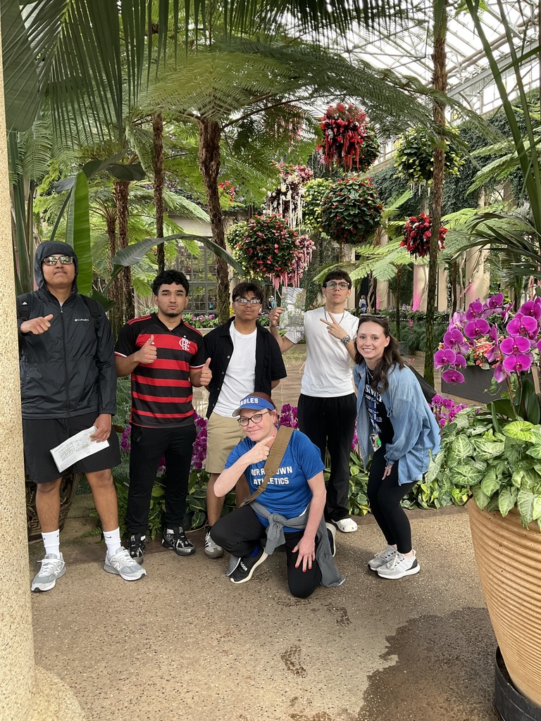 Students and teachers are posing together inside of the greenhouse next to flowers