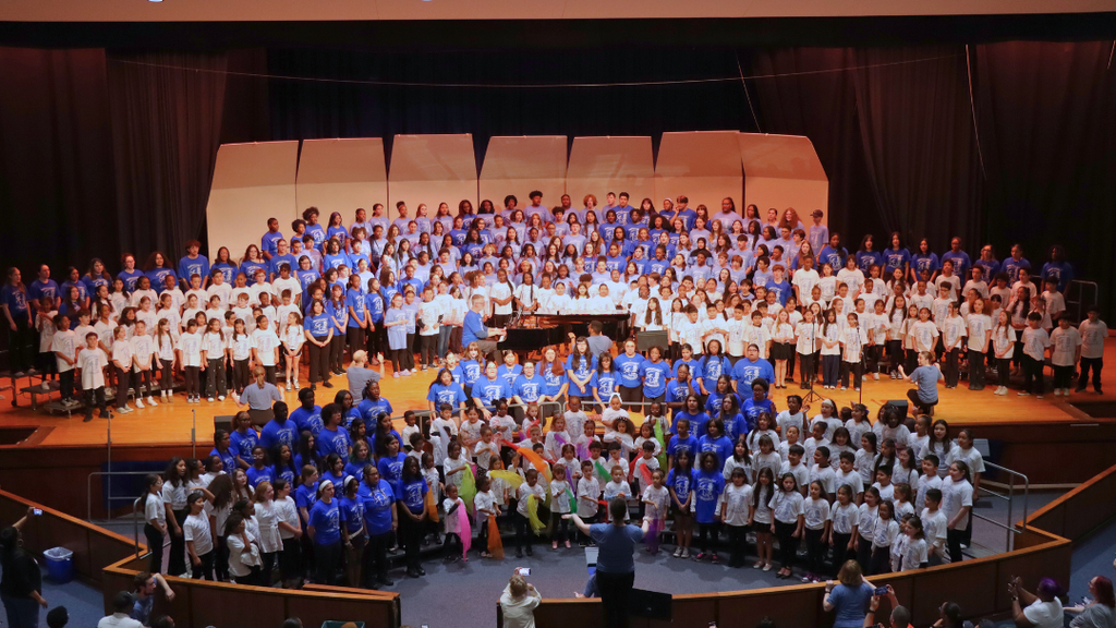 A large student choir in blue and white shirts performs on stage in an auditorium.