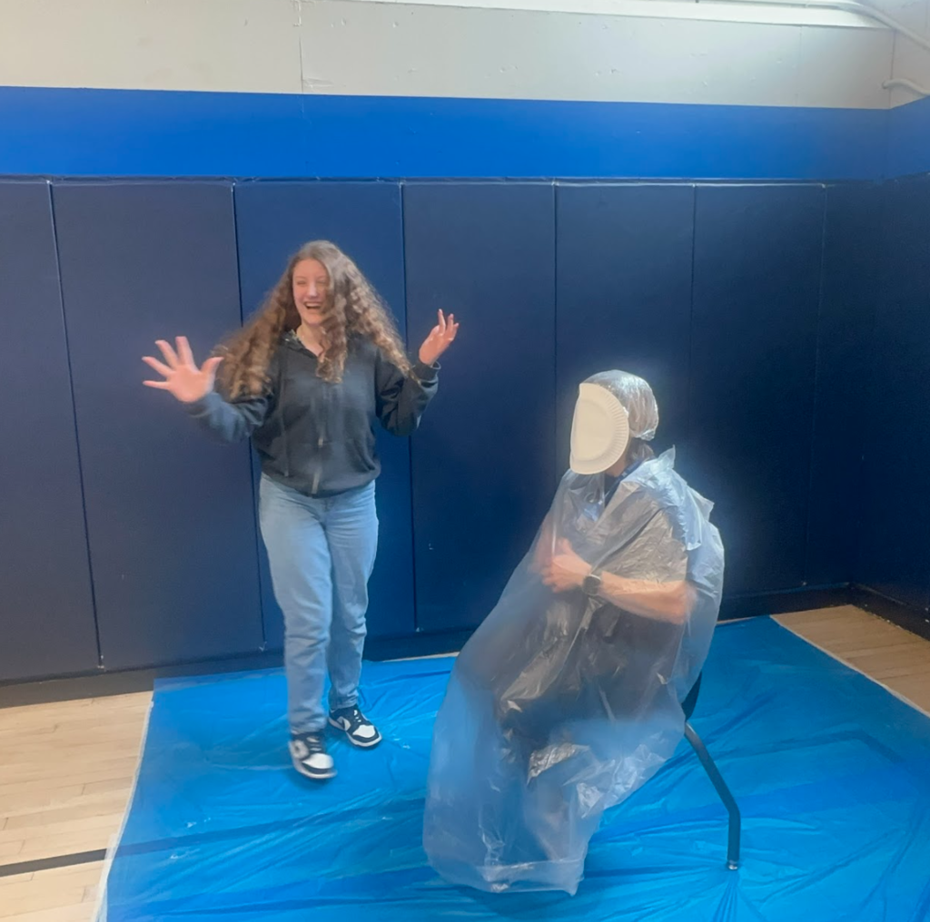 A student has her arms up and laughing and a teacher has a pie in their face