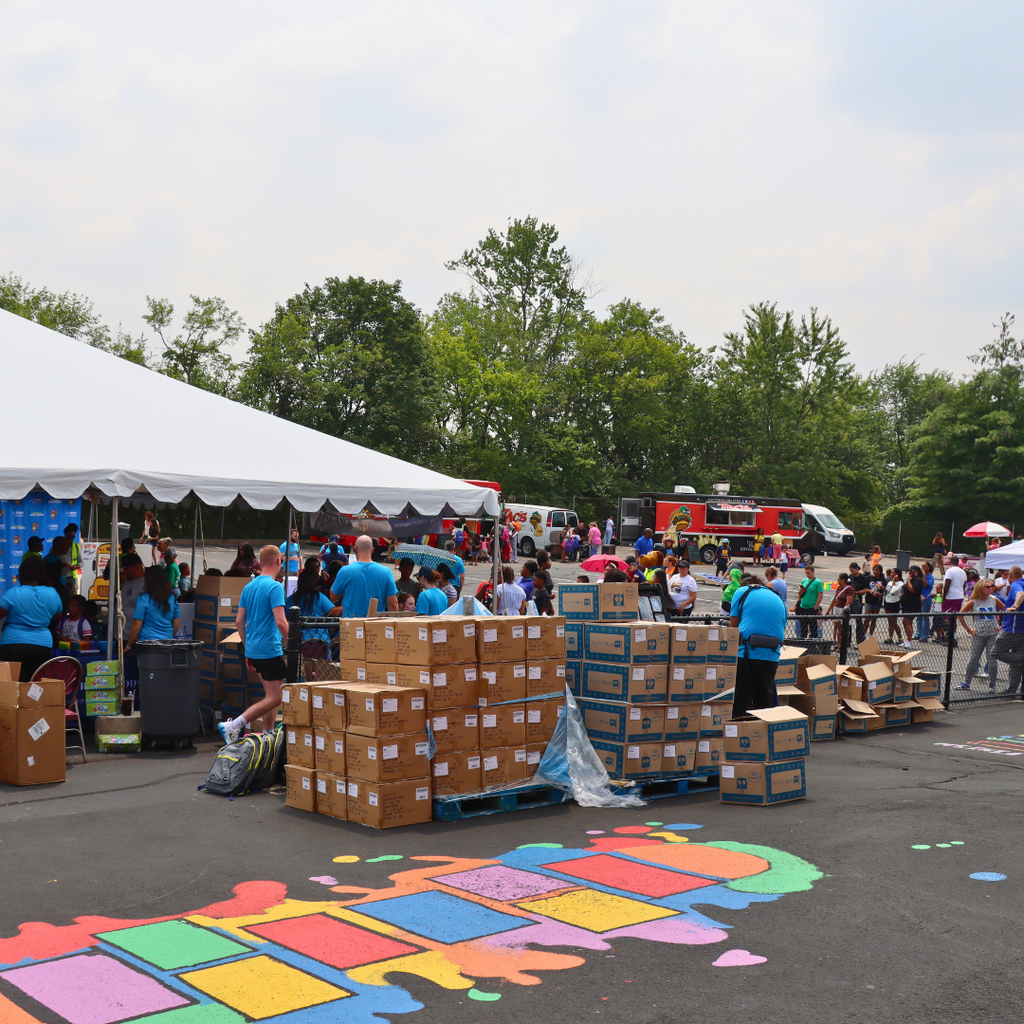 Project backpack event with people in blue shirts organizing boxes, a white tent, a painted hopscotch pattern, and food trucks in the background.