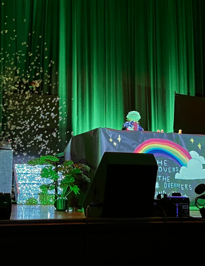 A student is using a kermit the frog puppet for his talent portion. Bubbles are also coming out behind him.