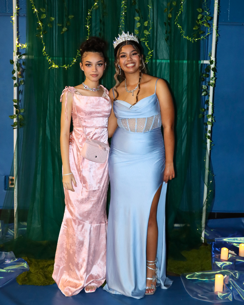 Two students at the junior prom posing for photo in front of green backdrop decorated with vines.