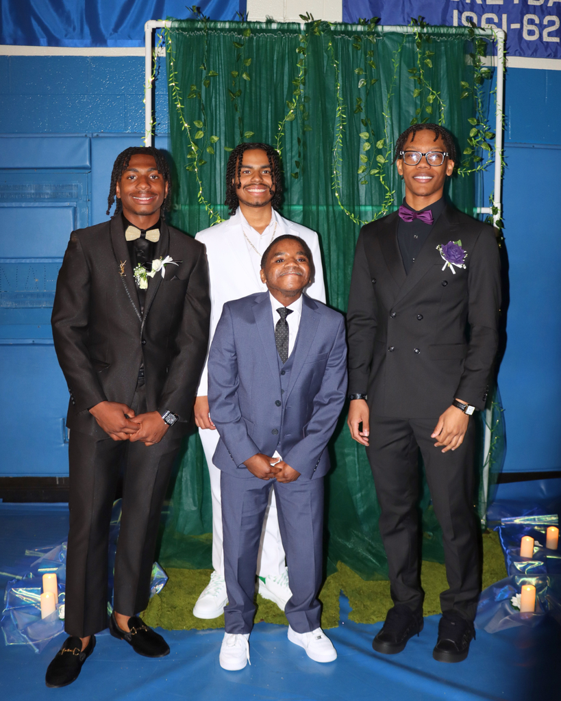 Four students at the junior prom posing for photo in front of green backdrop decorated with vines.