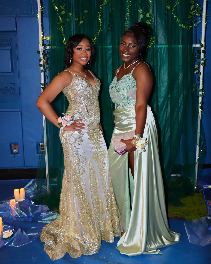 Two students at the junior prom posing for photo in front of green backdrop decorated with vines.