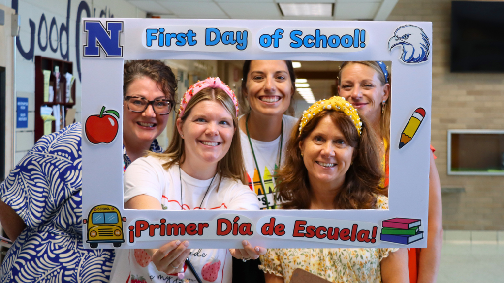Group of five teachers pose for photo with poster board frame that says First Day of School.