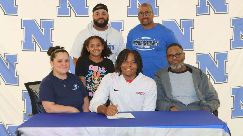 Senior student-athlete sitting at signing table with family and coaches.