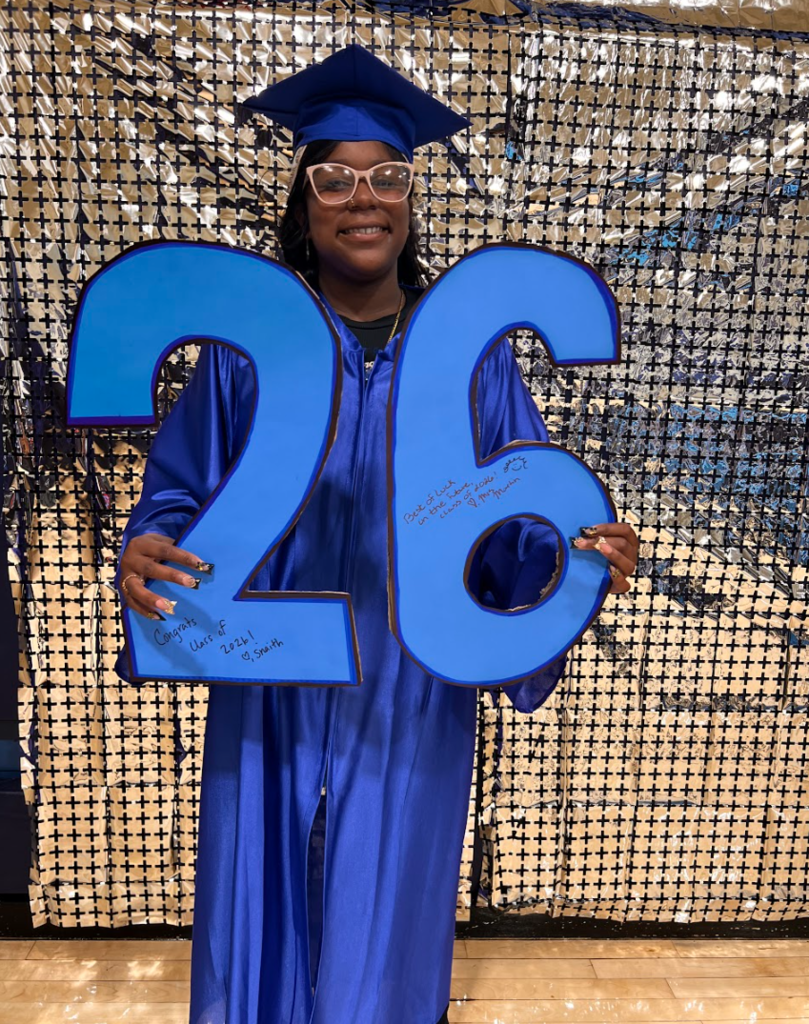 A student is holding a class of 26 sign in her cap and gown