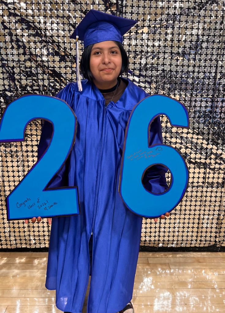 A student is holding a class of 26 sign in her cap and gown