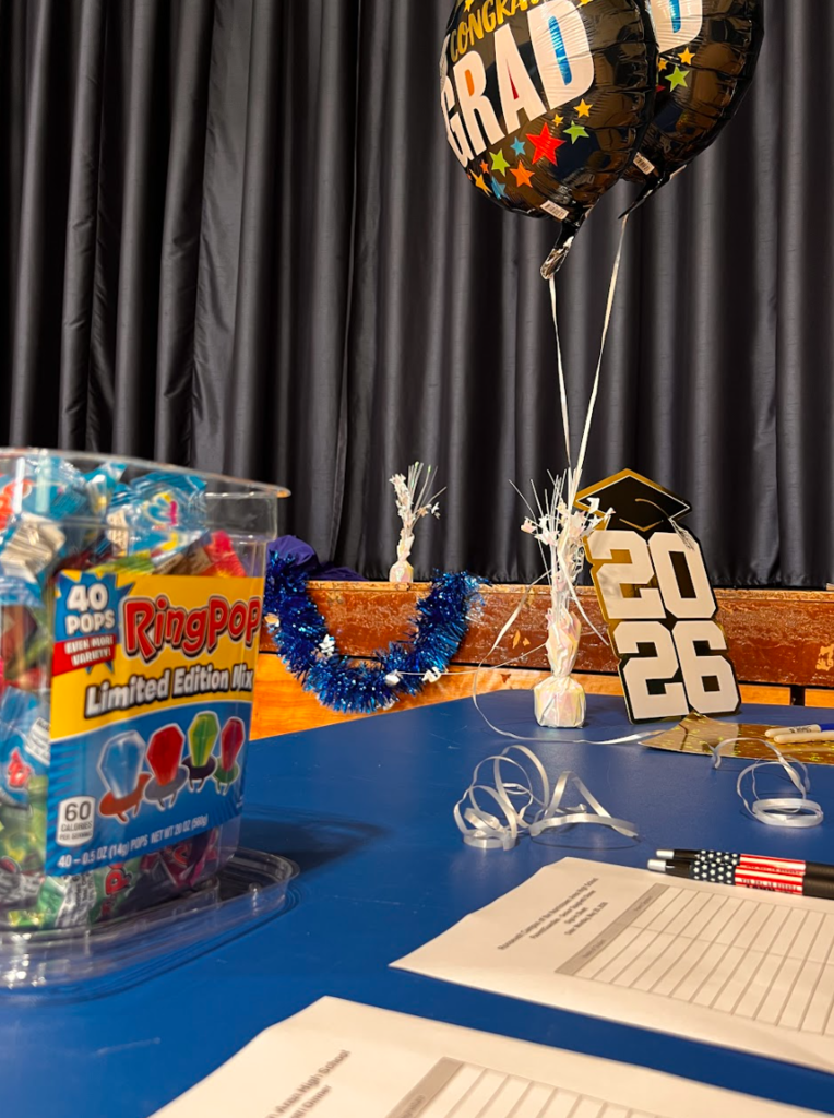 The table has ring pops and a class of 2026 sign as families come into the gym