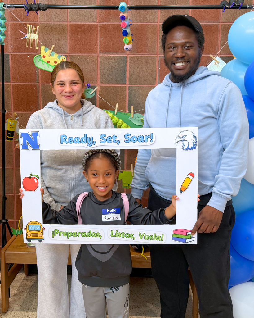 Student and their parents pose for a photo with a photo frame decorated with school items and text "Ready, Set, Soar!"
