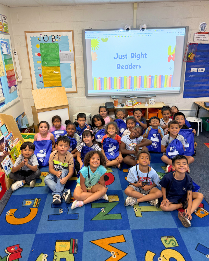 Students sitting on class rug with their packs of books, on the whiteboard says "Just Right Readers".