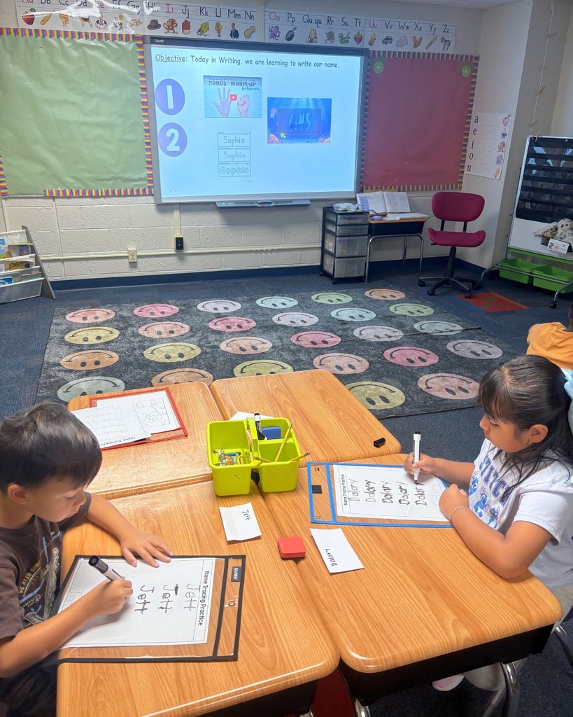 Student practicing writing their names on a whiteboard with instructions on the screen at the front of the room.