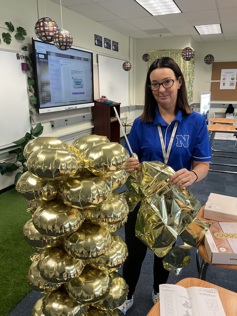 A teacher is setting up a balloon arch for her classroom and smiling at the camera.