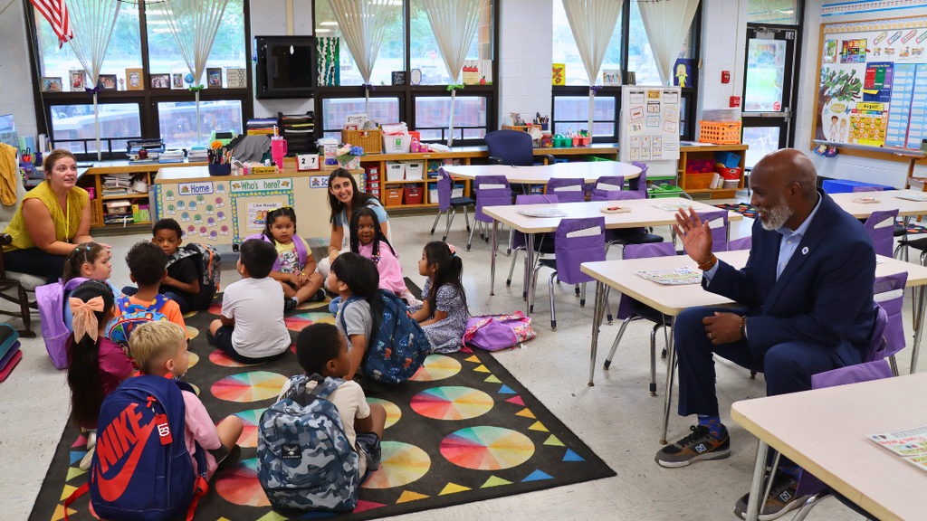 Dr. Green waving to new kindergarten students as they sit on the classroom carpet.
