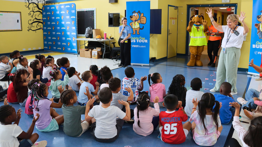 Students seated on the floor raise their hands during a safety event featuring costumed mascots and a “Safety Bus Tour” banner.