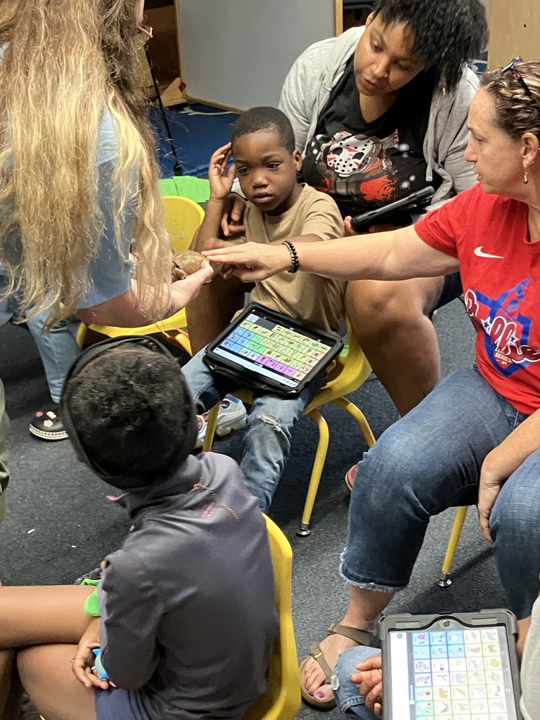 multiple adults and students look at and touch a small turtle as another adult holds the turtle out towards a student holding an AAC device