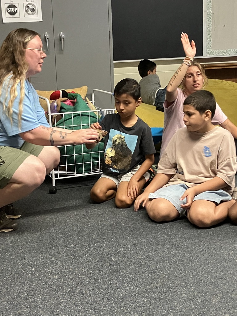a teacher sits near two boys on a rug as they look at and gently touch a small turtle held by a zookeeper