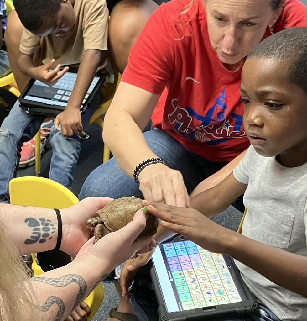 an A-Team student holding an AAC device touches a turtle