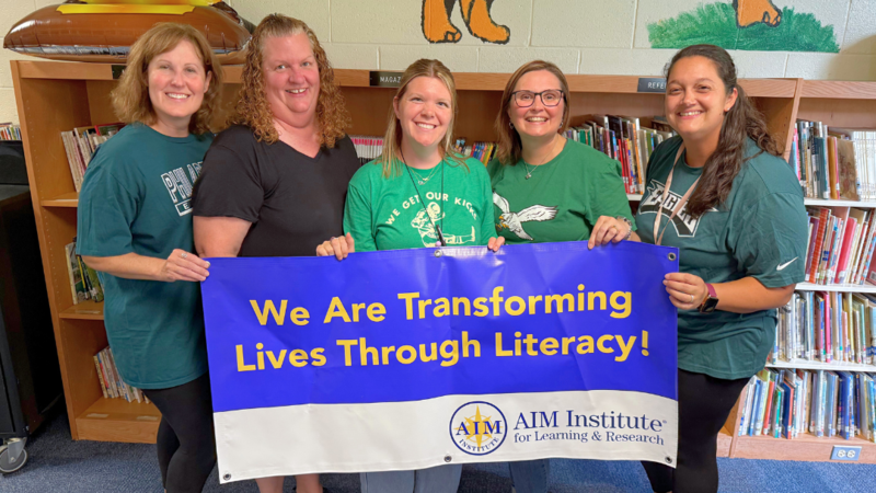 Marshall Street Staff pose for photo with AIM Institute Banner that says "We are transforming lives through literacy."