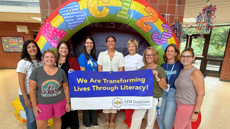Whitehall Staff pose for photo with AIM Institute Banner that says "We are transforming lives through literacy."