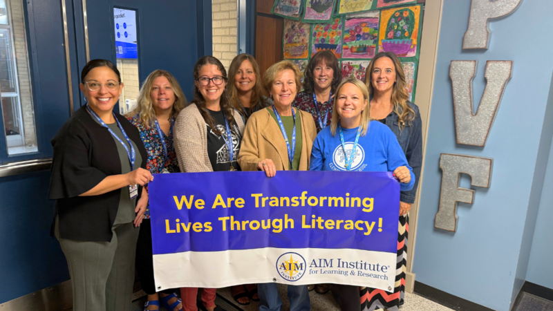 Paul Fly Staff pose for photo with AIM Institute Banner that says "We are transforming lives through literacy."