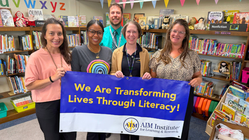 Cole Manor Staff pose for photo with AIM Institute Banner that says "We are transforming lives through literacy."