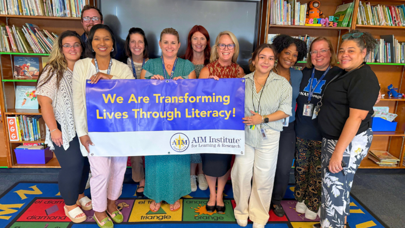 Hancock Staff pose for photo with AIM Institute Banner that says "We are transforming lives through literacy."