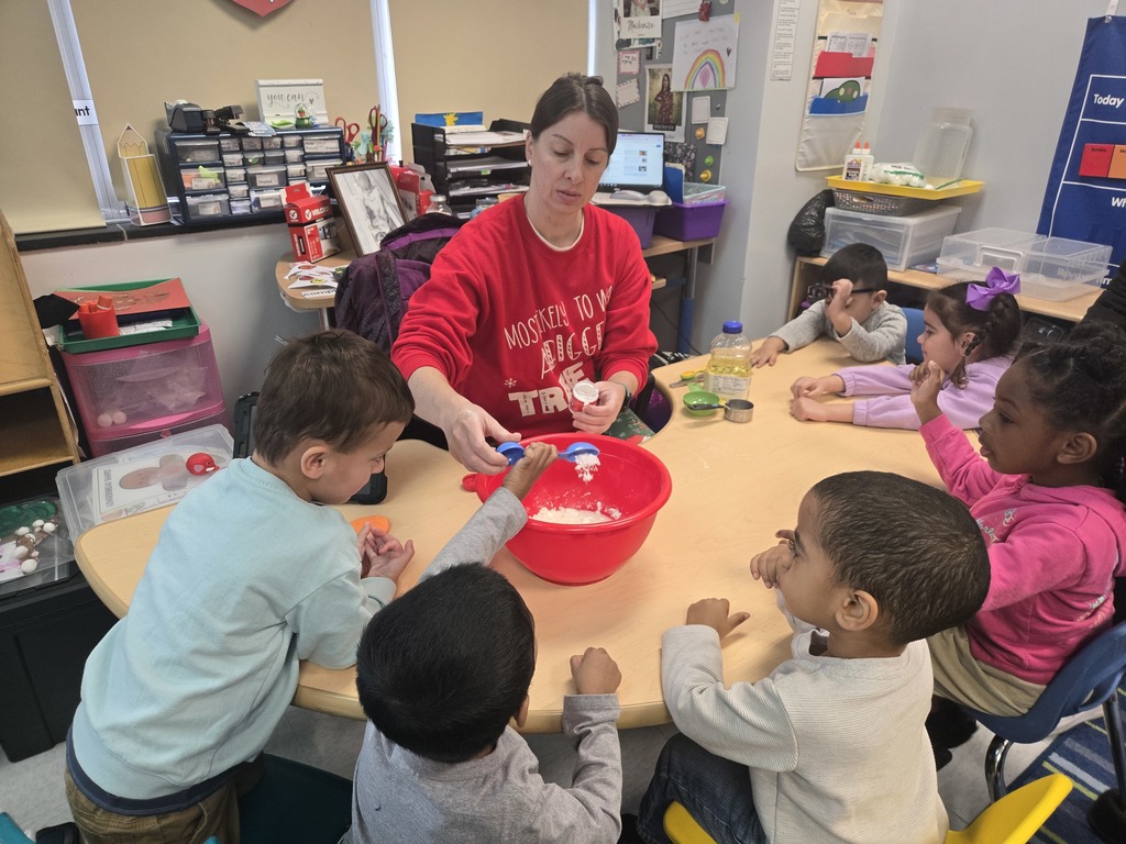 A preschool student is assisted in pouring ingredients into a red bowl while creating peppermint playdough