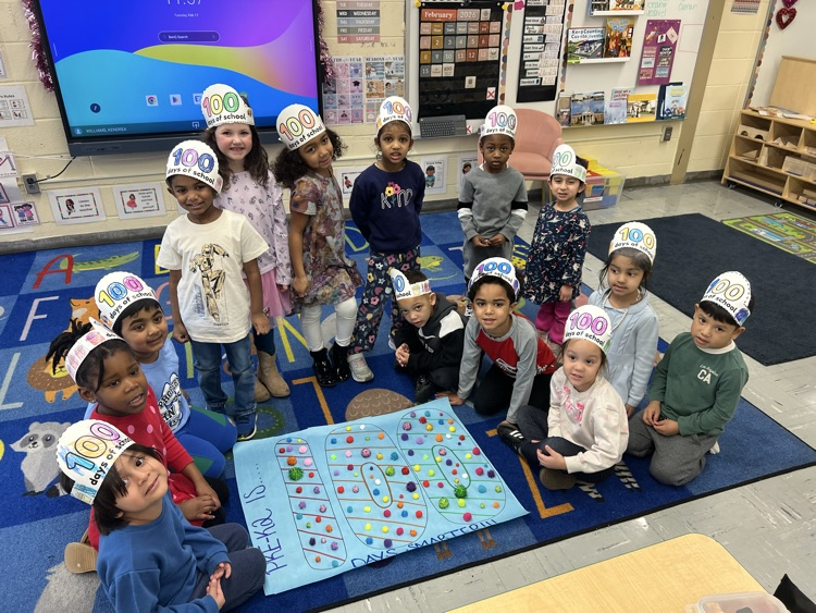 Group of elementary aged students in a classroom with hats that say 100 days standing in front of a  poster with pom poms on it