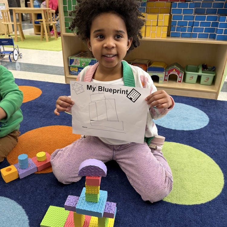 Student building with shapes on the floor and holding her blueprint