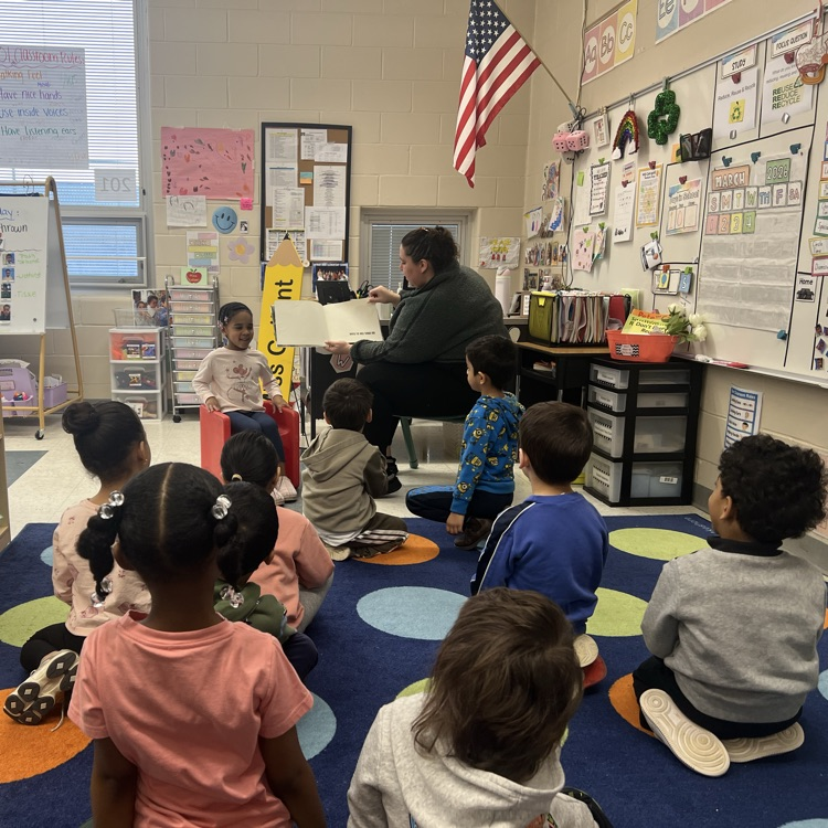 Students listening to an adult read a book in a classroom