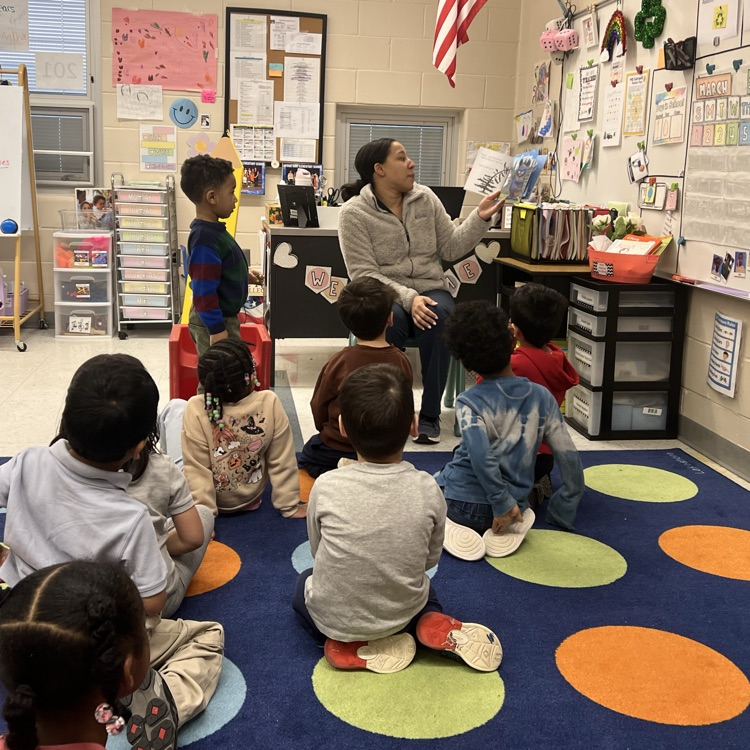 Students listening to an adult read a book in a classroom