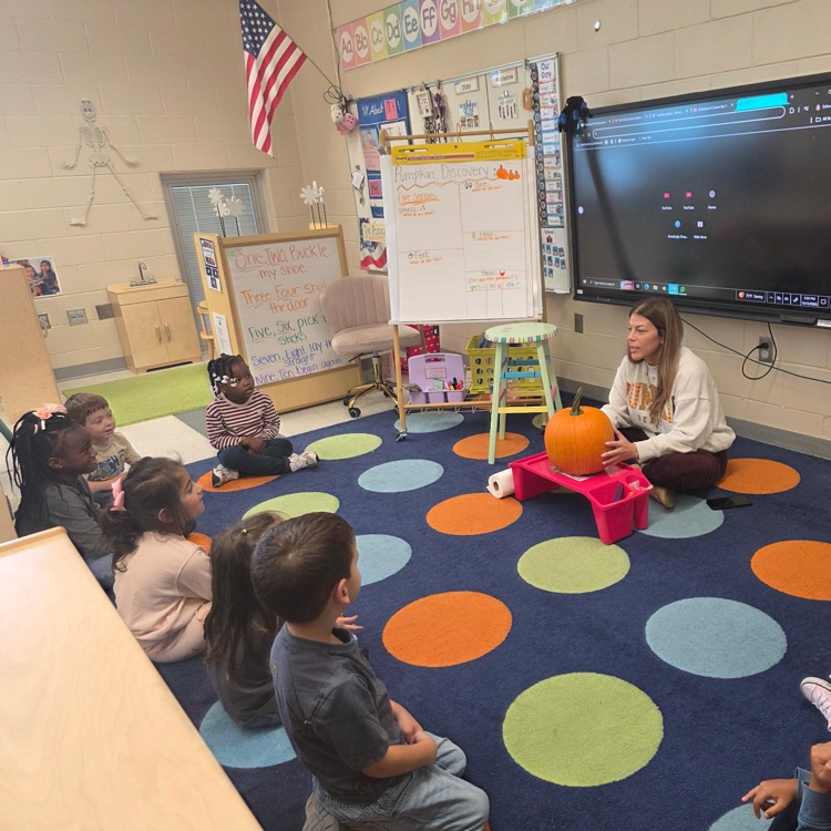Teacher holding a pumpkin and students listening to her on the carpet
