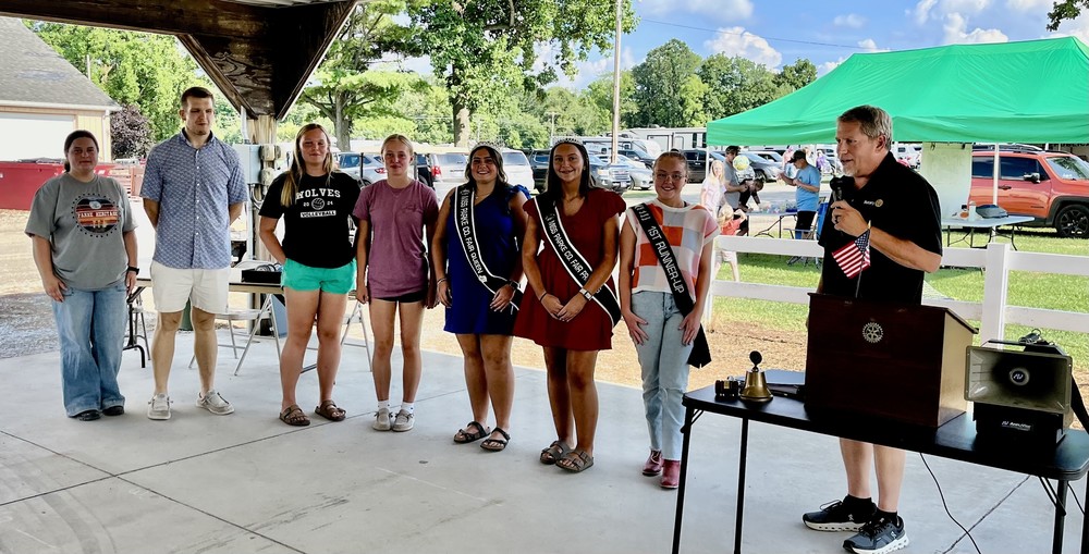 eight people standing in the Pavilion at the fairgrounds with one person talking and recognizing the other seven