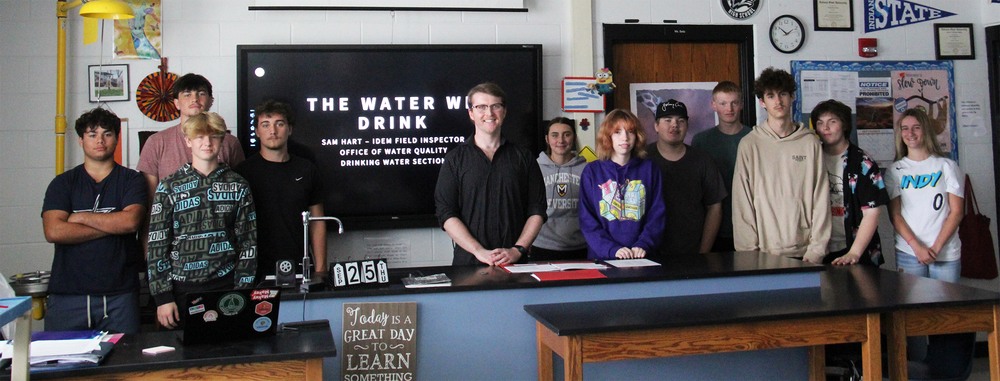 students along with the guest speaker standing in a row behind a science desk