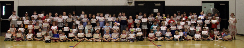 students sitting or standing on the gym floor holding certificates for perfect attendance