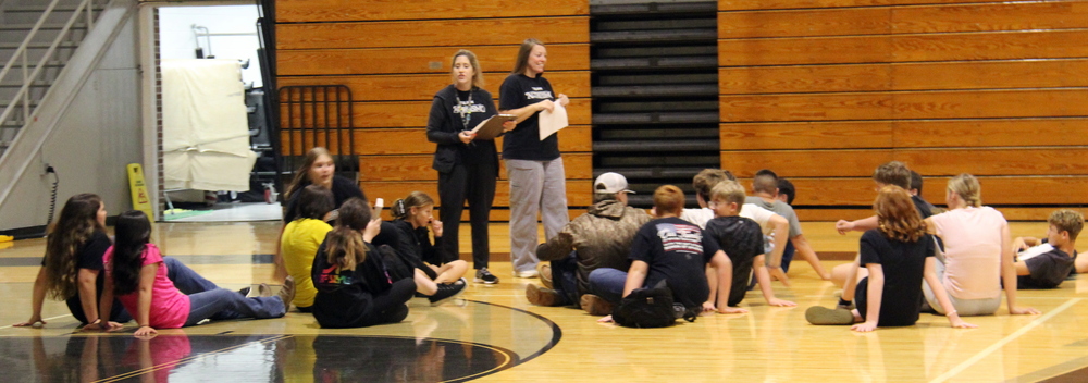 students sitting on the floor of the gym and two adults standing in the middle