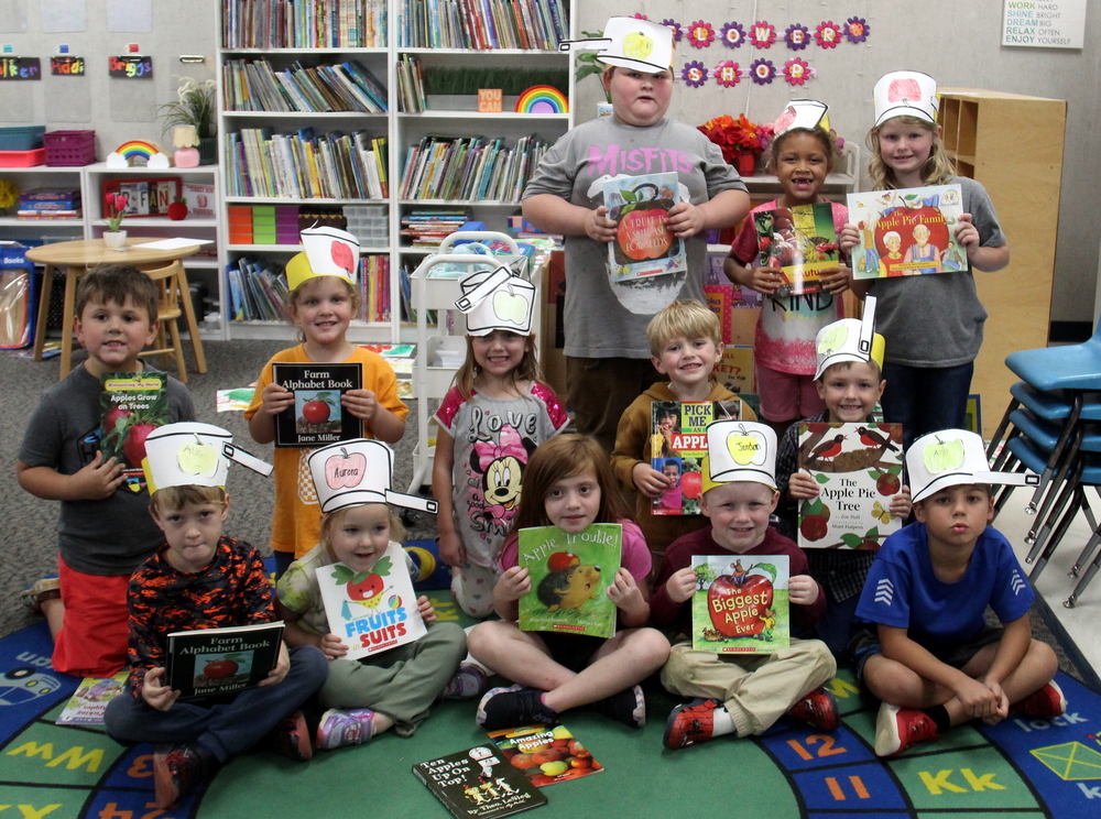 students sitting and standing on a carpet wearing Johnny Appleseed hats and holding books about apples