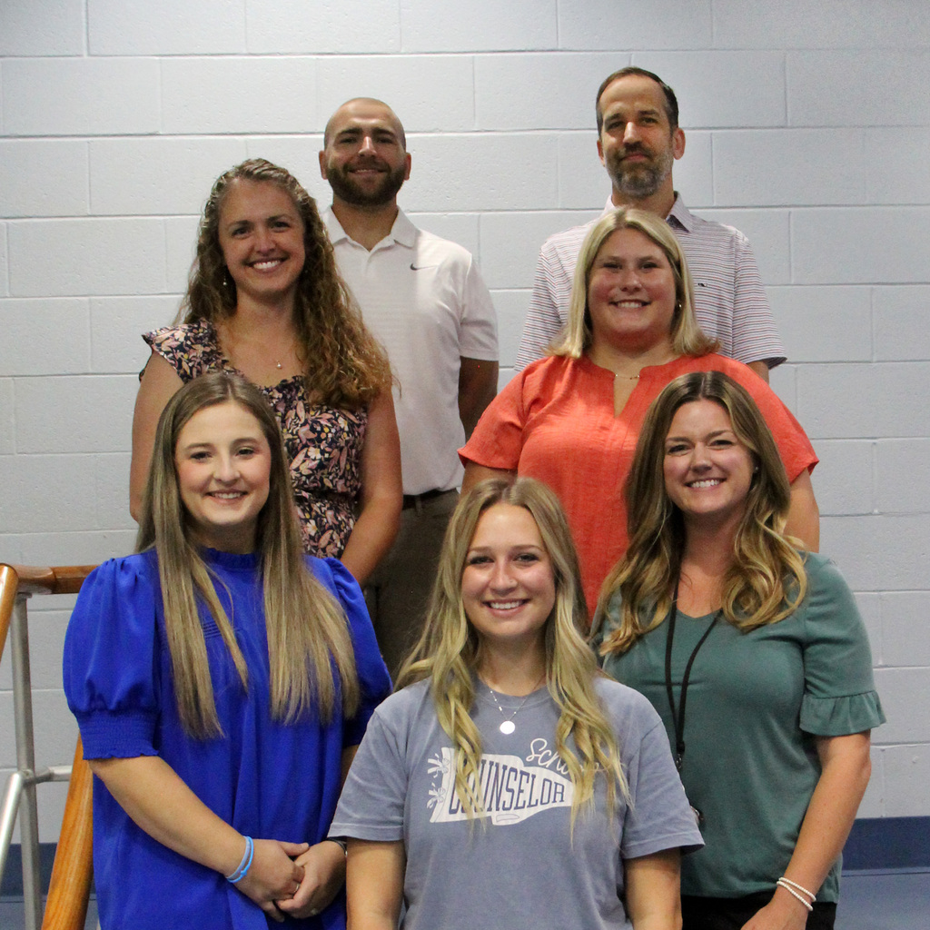 Group photo of seven new smiling staff members standing in three rows indoors against a white brick wall.