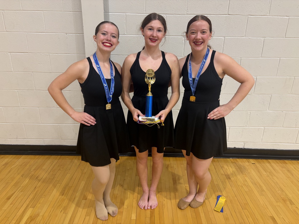Two dance girls with medals around their neck and 1 dance girl in the middle holding a trophy, smiling for a photo.