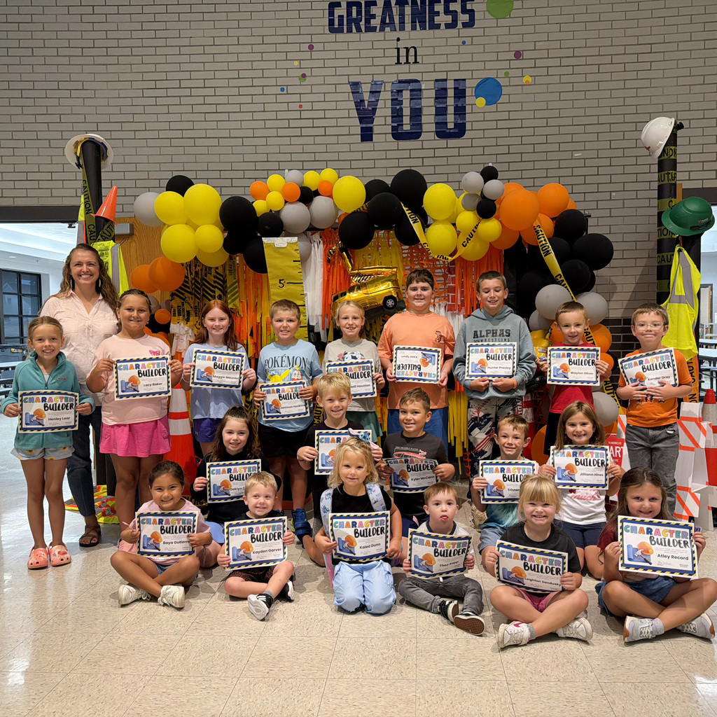 A group of elementary school students pose with an adult in a school hallway. The students are arranged in three rows and are smiling while holding certificates that read “Character Builder.” Behind them is a colorful construction-themed display with yellow, black, orange, white, and blue balloons, caution tape, construction cones, and decorations.