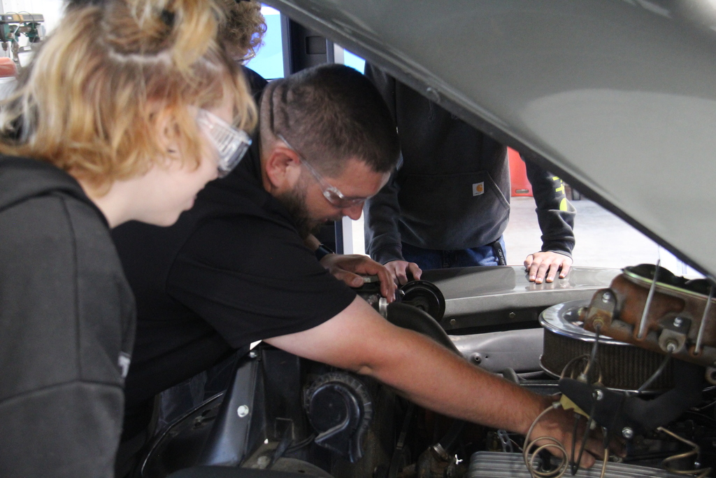 Auto Service teacher leans under the hood of a car to show 2 students something that could be wrong with the vehicle. 